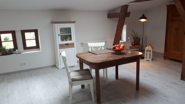 Ansicht des Essbereichs mit Holztisch, darauf eine Schale mit Äpfeln, zwei Stühlen, im Hintergrund zwei kleine Fenster, ein kleiner weißer Schrank, ein Kleiderschrank aus Holz